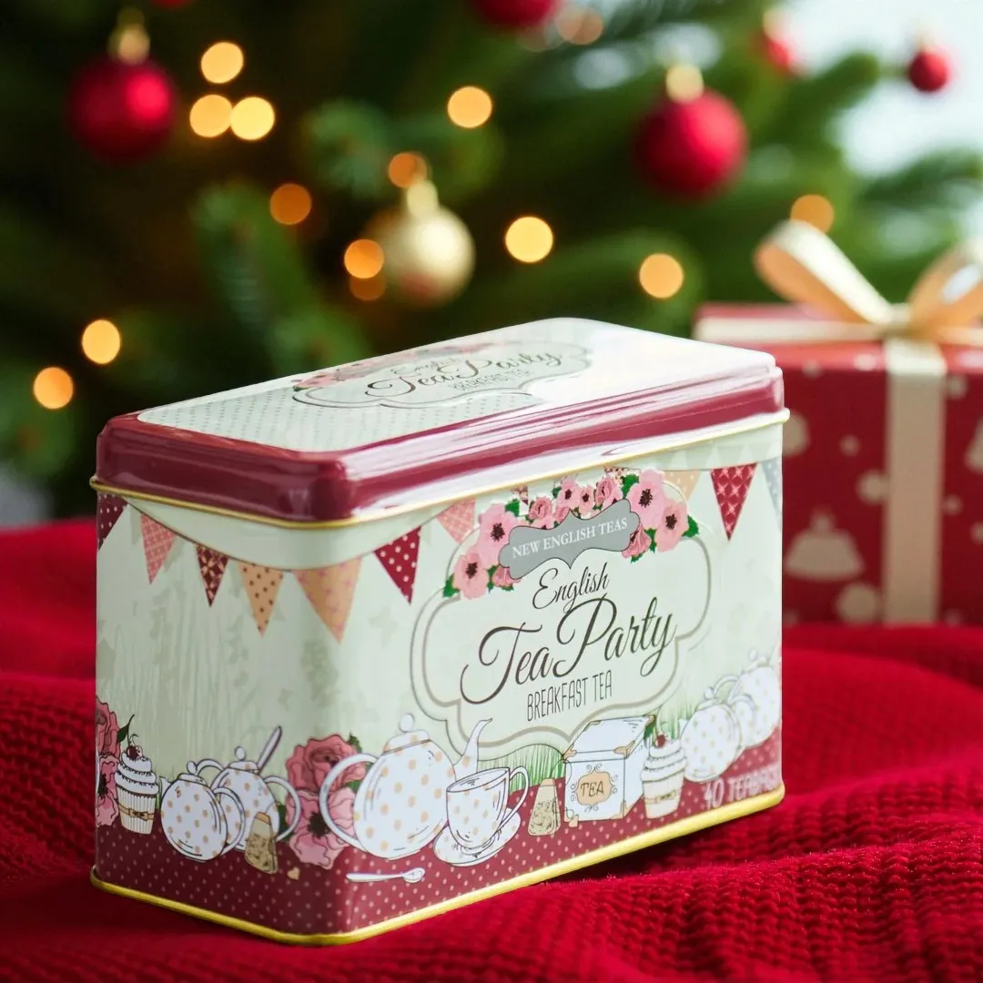 An English Tea Party Red 40 Teabag Tin sits on a red knit blanket in front of a Christmas tree decorated with red and gold ornaments.