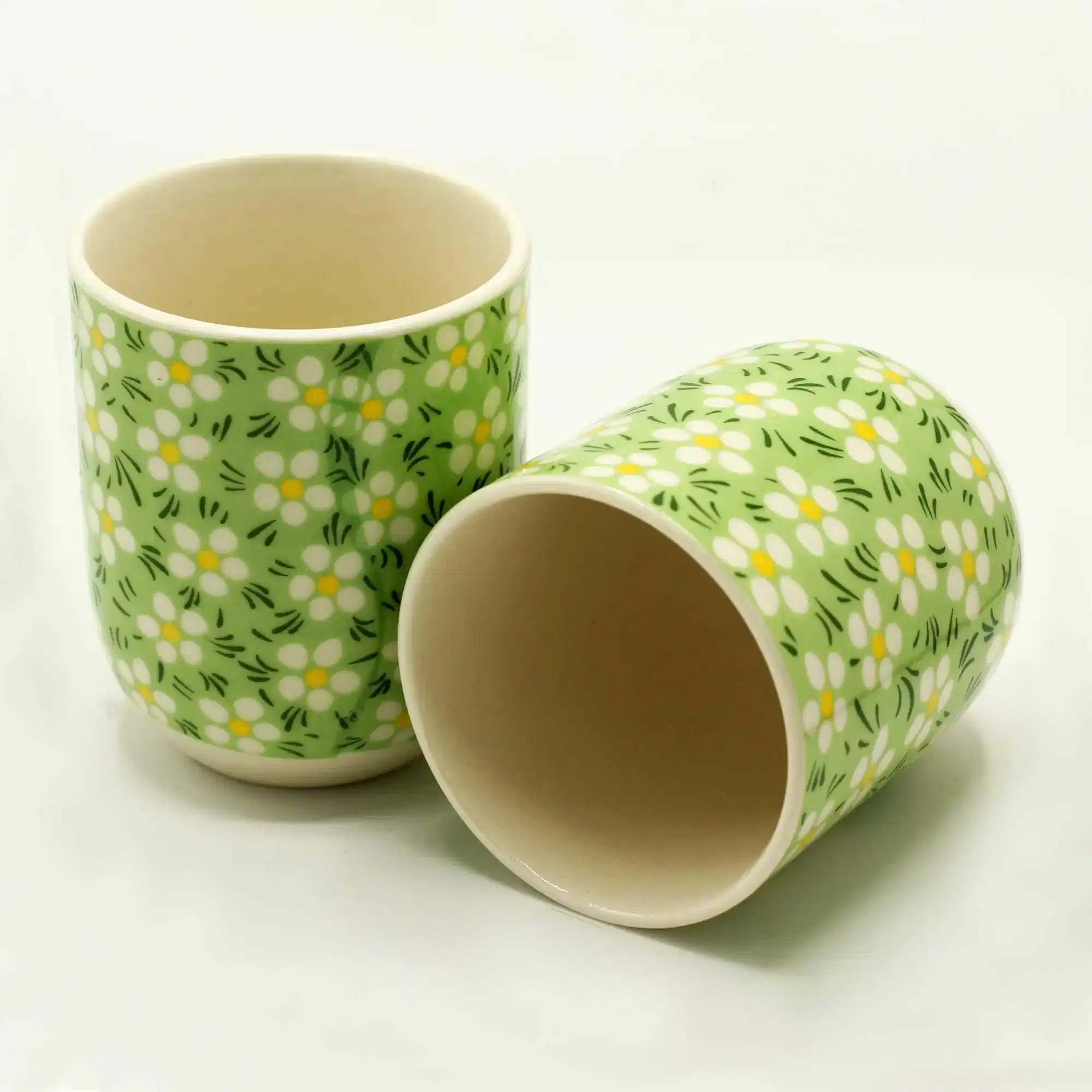 Two ceramic cups with a green background and white daisy flower pattern, one standing upright and the other lying on its side.