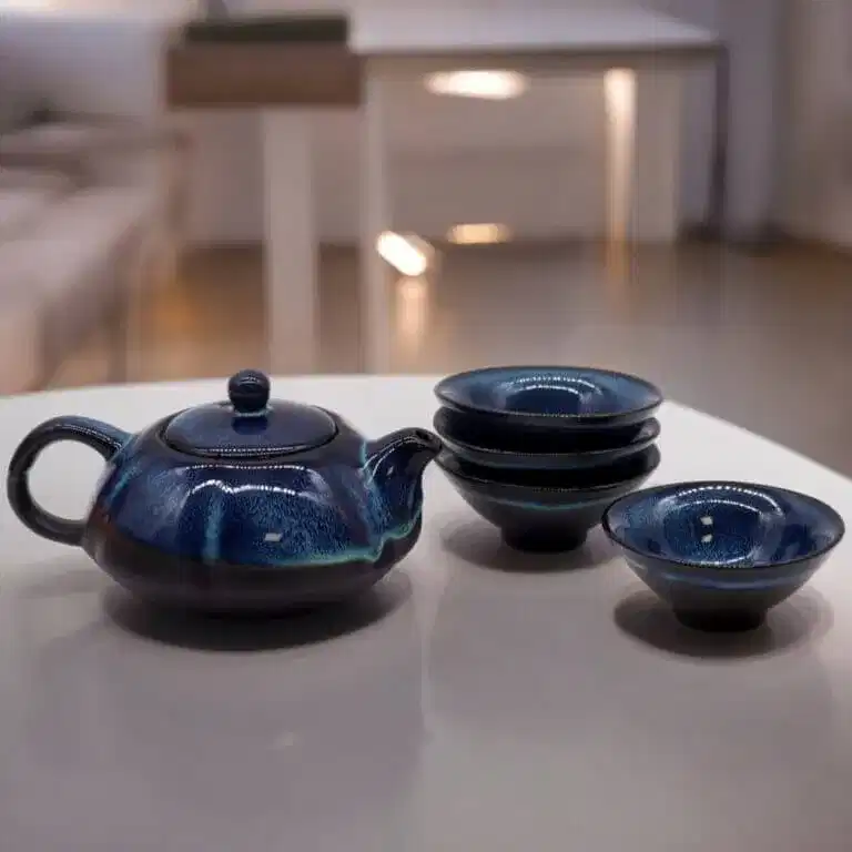 A dark blue ceramic teapot is placed on a white table next to four matching teacups, three of which are stacked. The background is softly lit.