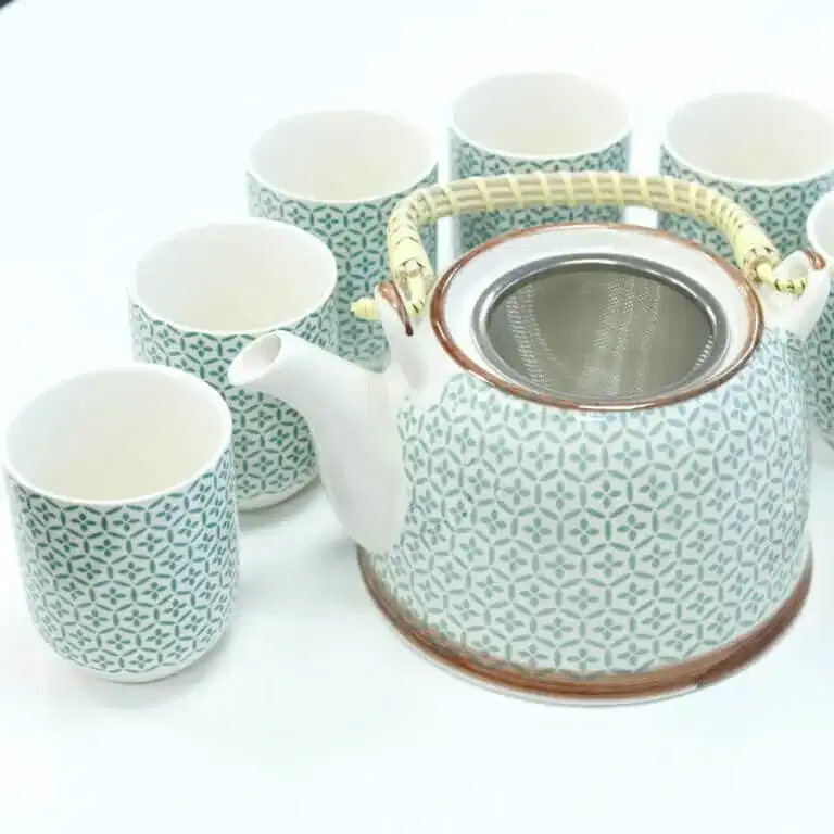 A patterned ceramic teapot with a wicker handle is surrounded by six matching teacups, all on a white surface.