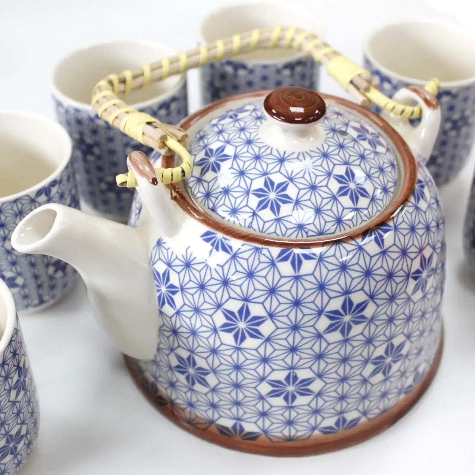 A ceramic teapot with a blue geometric pattern and woven handle is surrounded by matching teacups on a white surface.