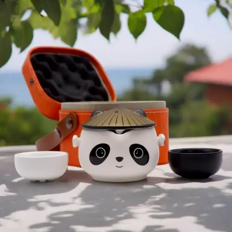 A panda-shaped teapot with a conical lid is displayed next to a white teacup, a black teacup, and an open orange carrying case outdoors.
