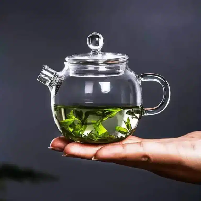 A Petite Glass Bubble Teapot with Lid, brimming with green tea leaves, is elegantly held in a person's hand against a dark backdrop.