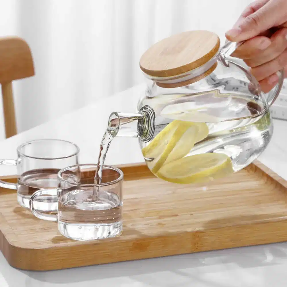 A Nordic Glass Teapot with a Bamboo Lid & Steel Spout gracefully pours lemon water into a glass on a wooden tray, next to an empty glass.