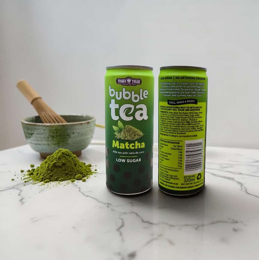 Two cans of Tiger Tiger bubble tea matcha flavor on a marble surface, with a bowl of matcha powder and a bamboo whisk in the background.