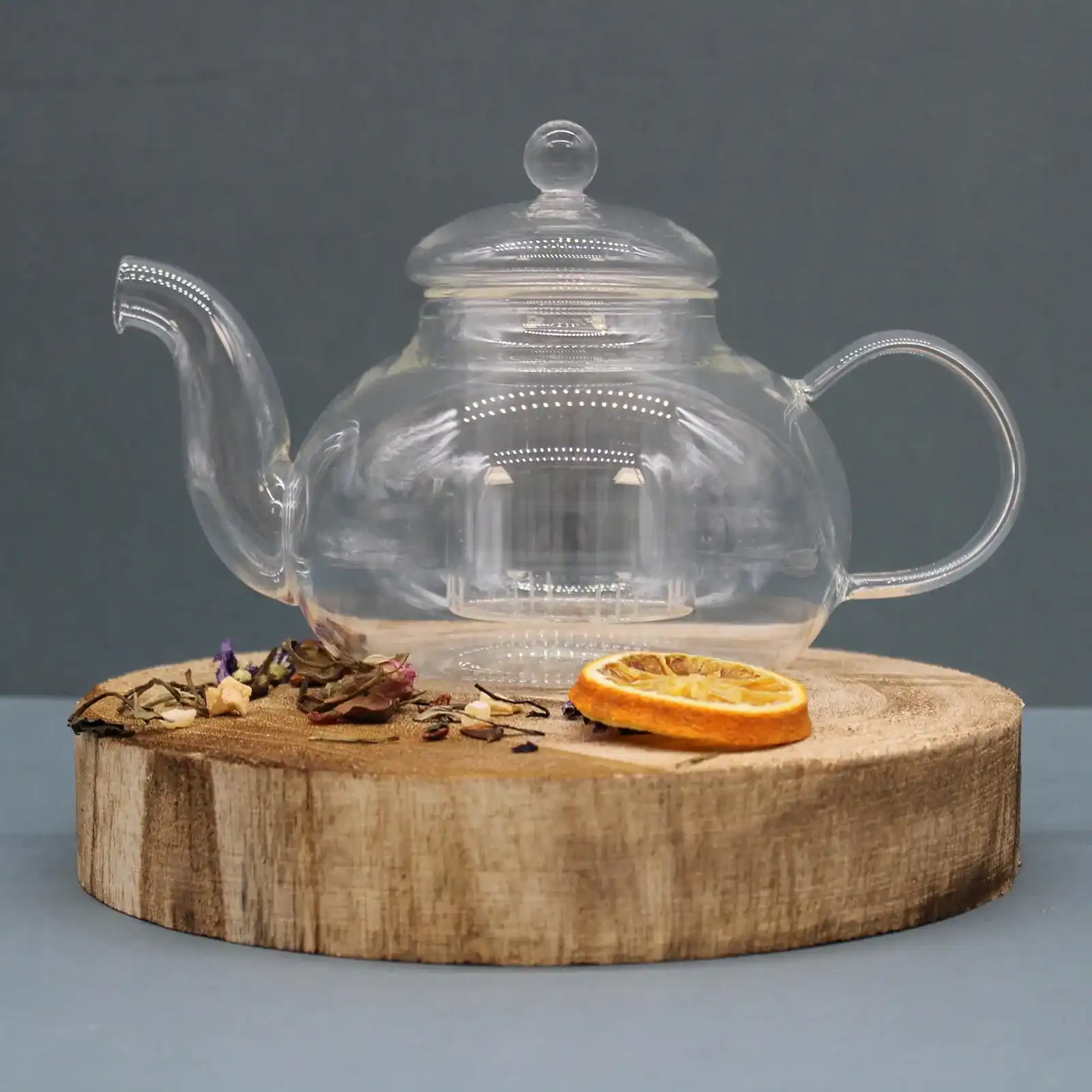A clear glass teapot sits on a round wooden board with dried tea leaves and a slice of dried orange beside it, against a plain gray background.