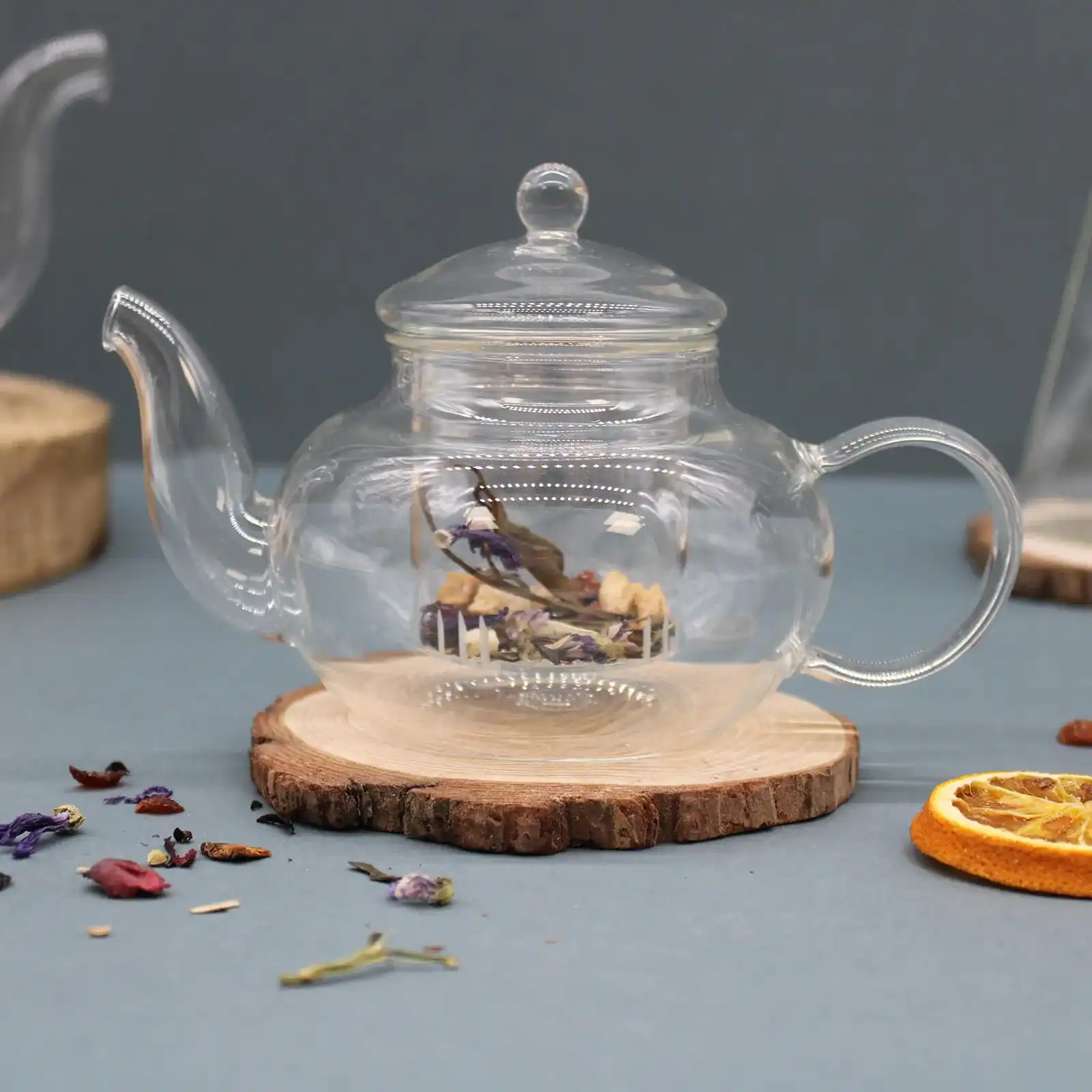 A clear glass teapot sits on a wooden coaster, containing loose dried herbs and flowers. Dried fruit and scattered tea leaves are nearby on a gray surface.