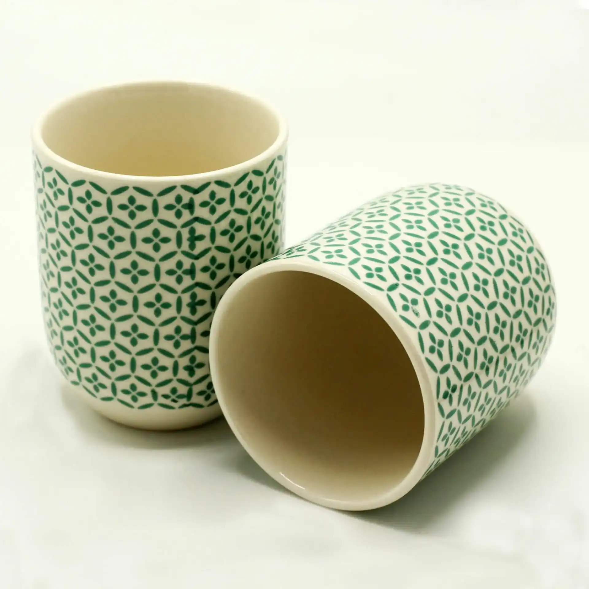 Two ceramic cups with a green geometric pattern, one standing upright and the other lying on its side, on a plain white background.