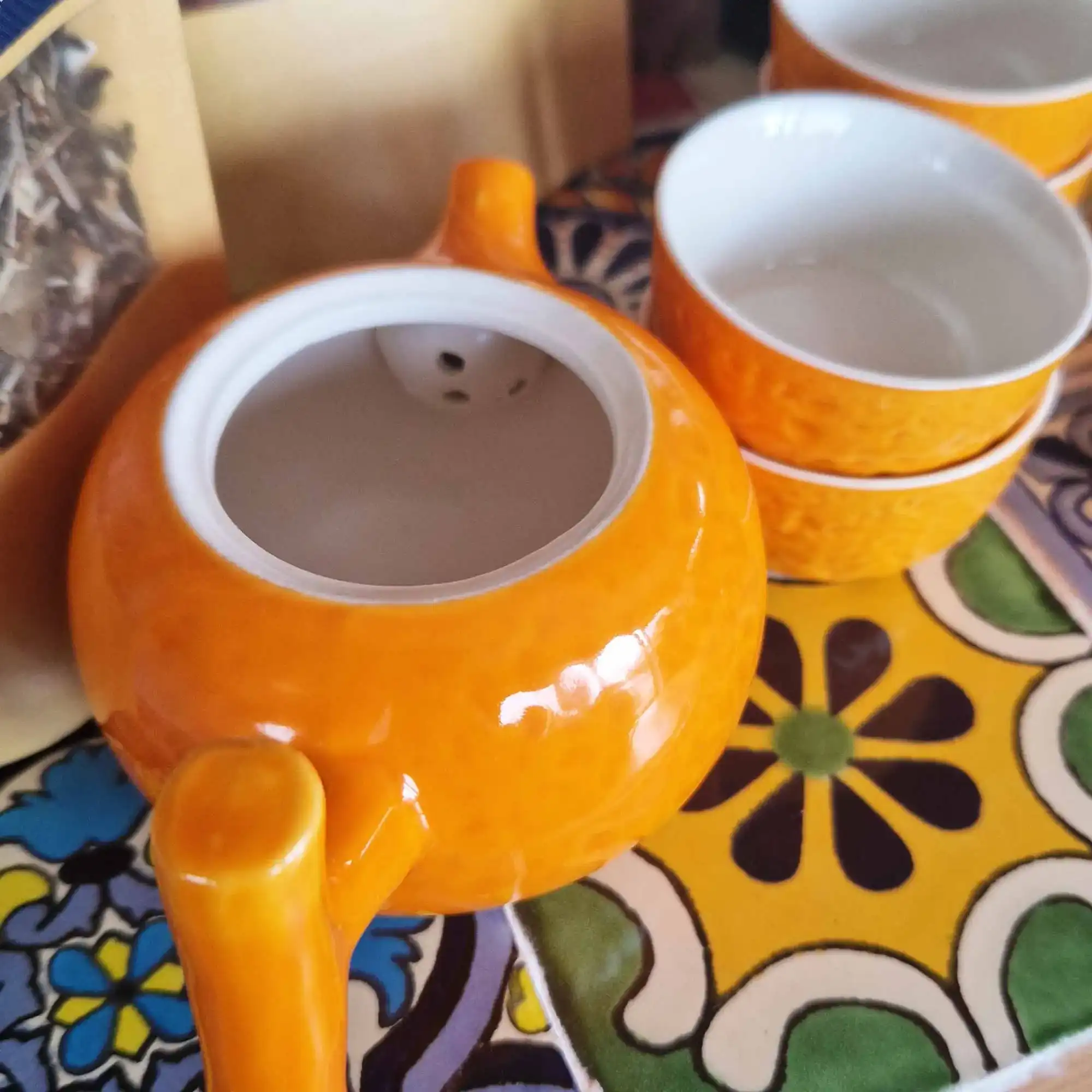 A bright orange teapot and two matching teacups are placed on a colorful, patterned surface with floral designs.