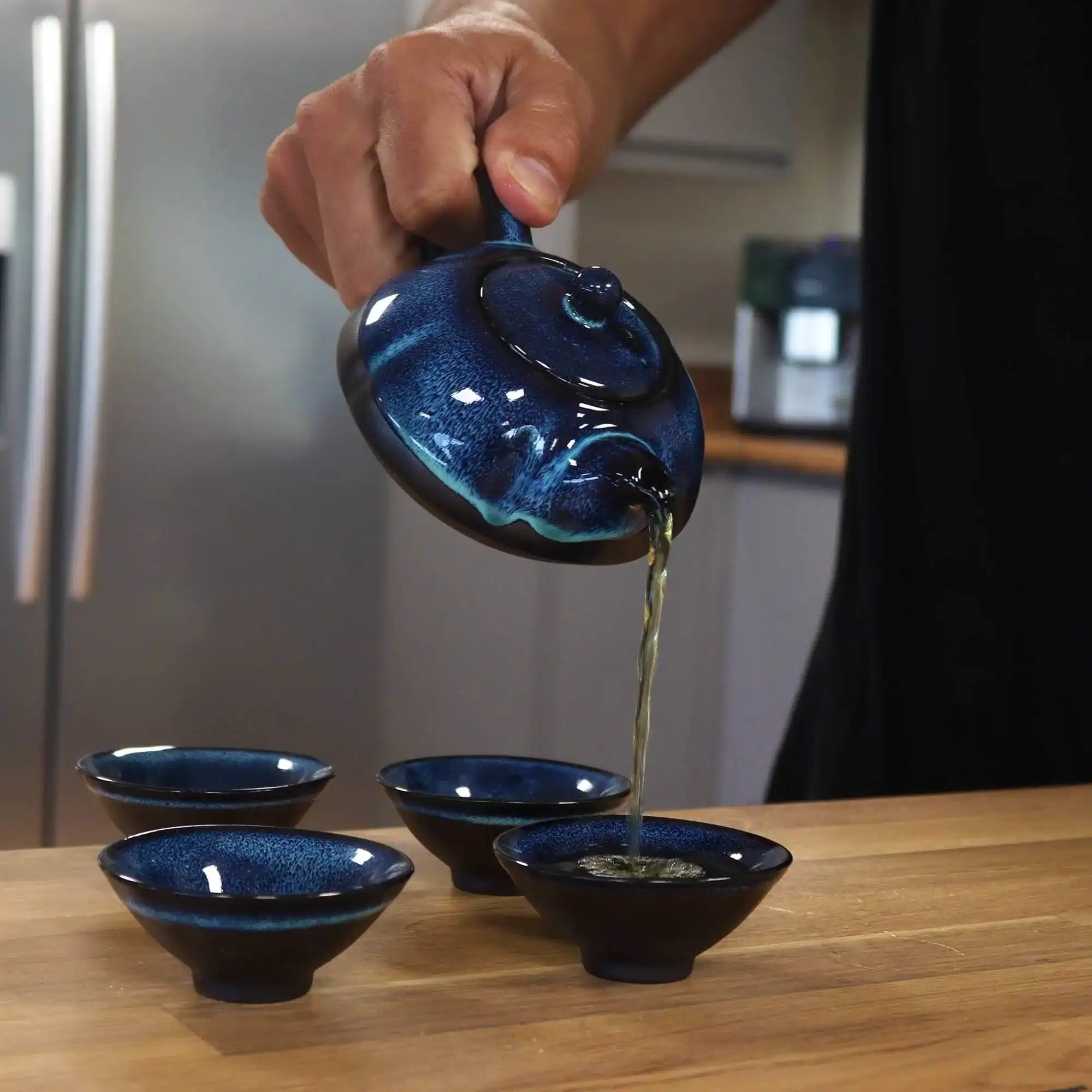 A hand pours tea from a blue teapot into one of four matching blue teacups on a wooden surface.