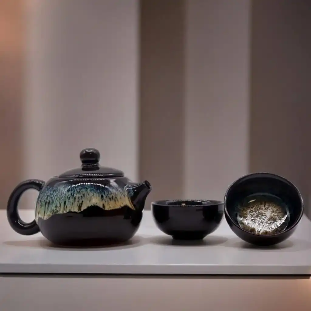 A black ceramic teapot with a textured glaze sits next to two matching teacups on a white surface against a softly blurred background.