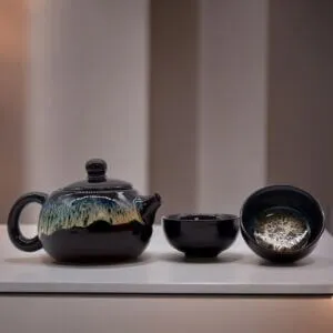 A black ceramic teapot with a textured glaze sits next to two matching teacups on a white surface against a softly blurred background.