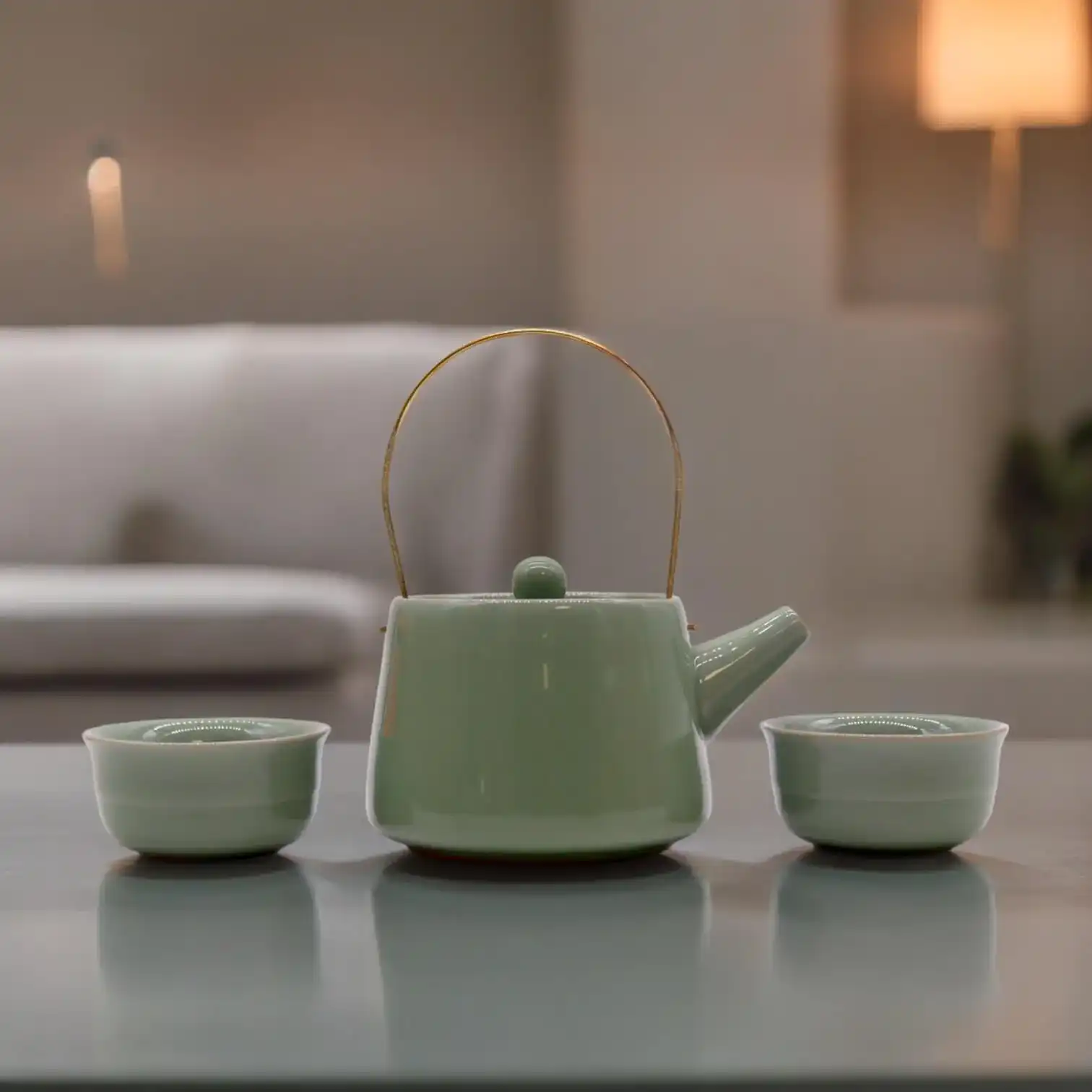 A ceramic teapot with a curved handle is placed between two matching teacups on a table, with a sofa and lamp blurred in the background.