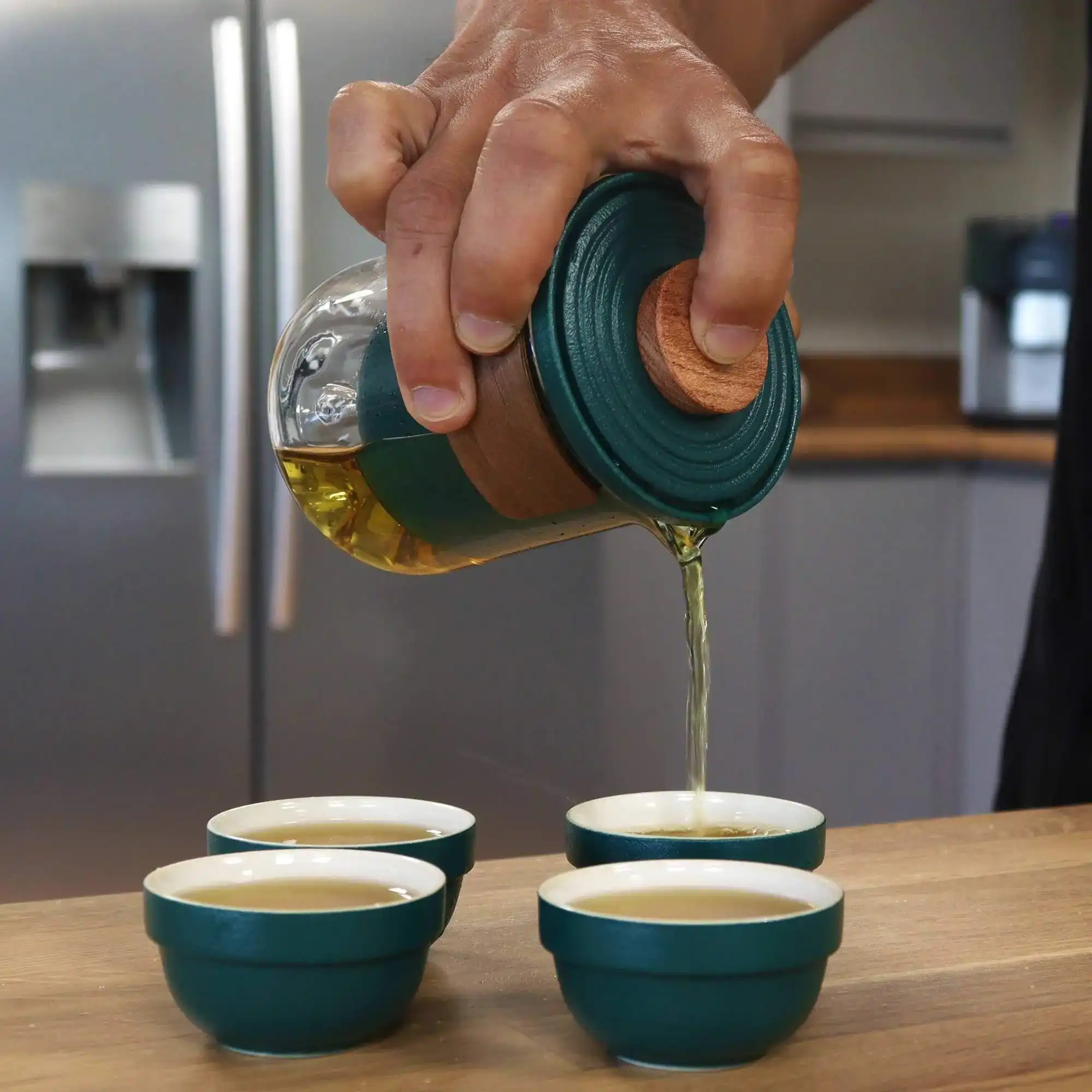 A hand pours tea from a glass teapot with a green lid into one of three small matching teacups on a wooden surface in a kitchen.