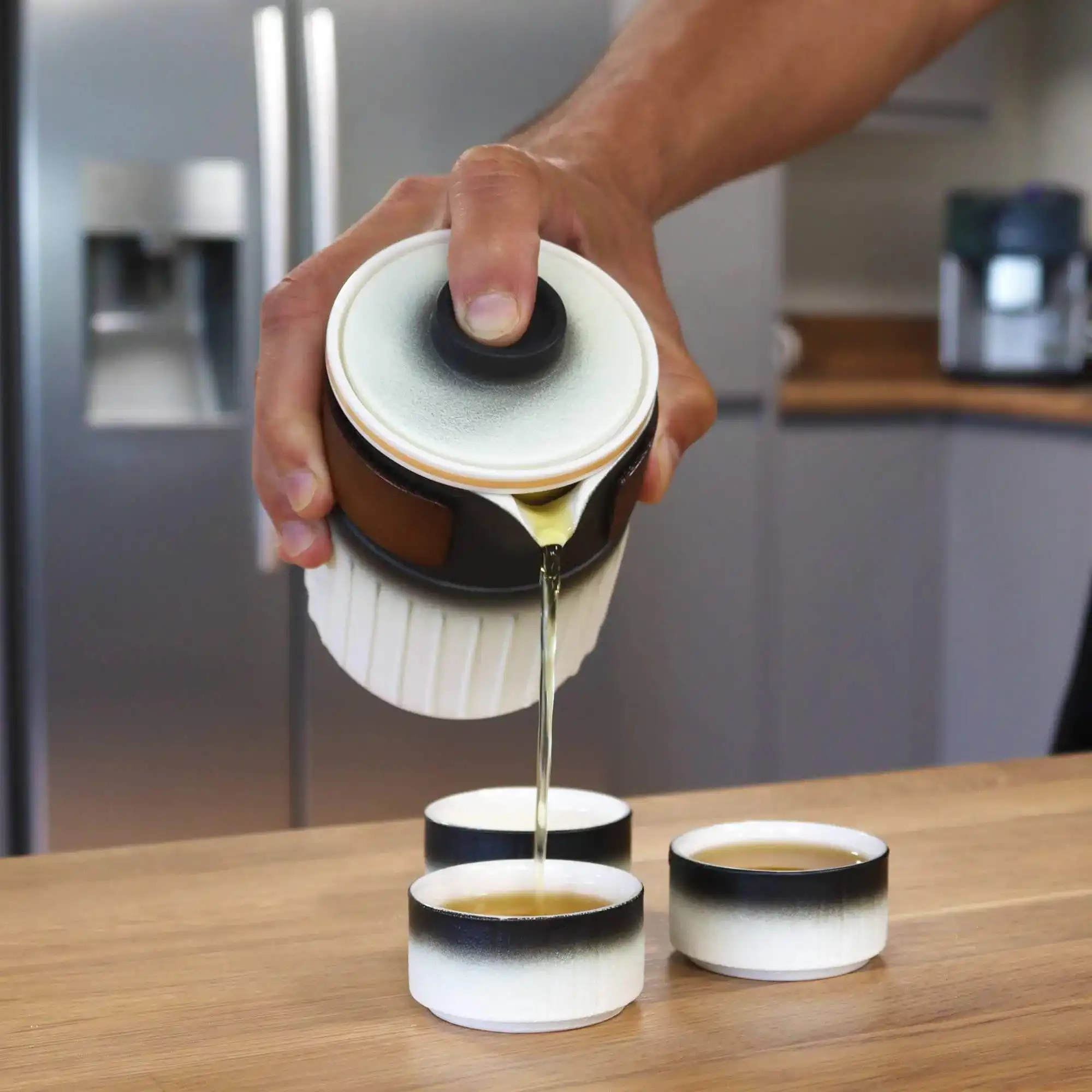 A person pours tea from a ceramic teapot into three matching teacups on a wooden countertop in a modern kitchen.