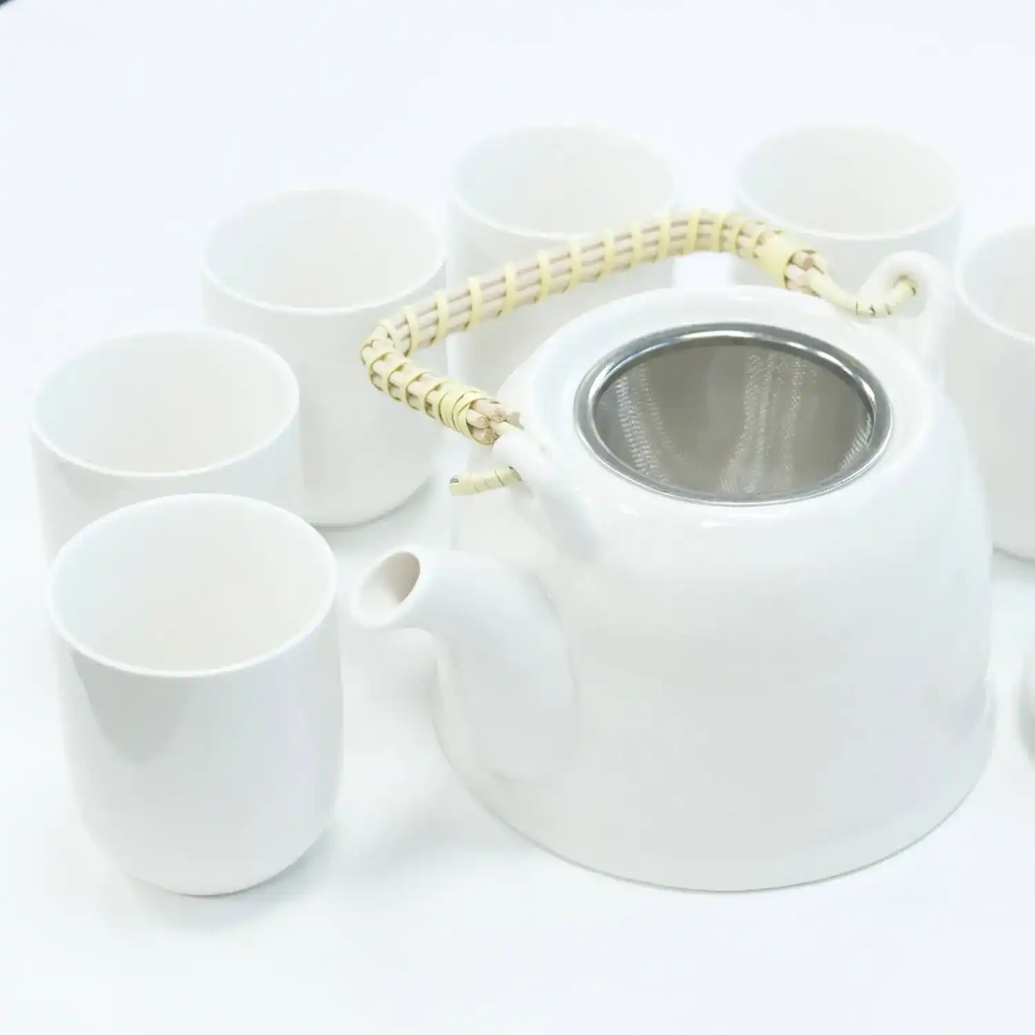 A white teapot with a woven handle and metal strainer is surrounded by six matching white cups on a white surface.