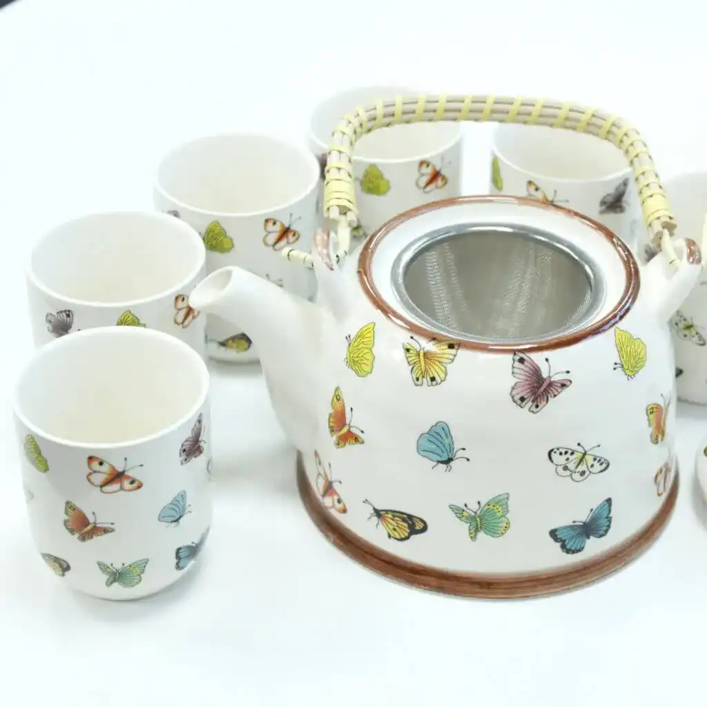 A ceramic teapot with a wicker handle and matching cups, all decorated with colorful butterfly illustrations, arranged on a white surface.