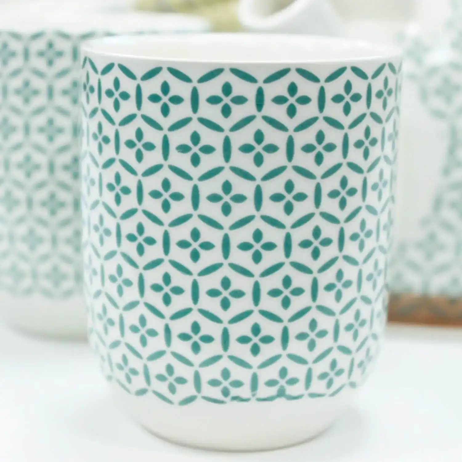 White ceramic cup with a teal geometric pattern consisting of repeating diamond and circle shapes. The cup is on a white surface, with another similar cup slightly blurred in the background.