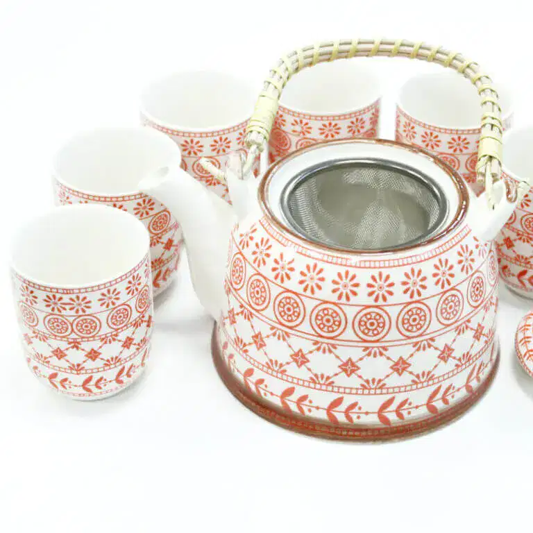 A ceramic teapot with a metal strainer and a bamboo handle, surrounded by five matching cups, all featuring a red and white geometric pattern.