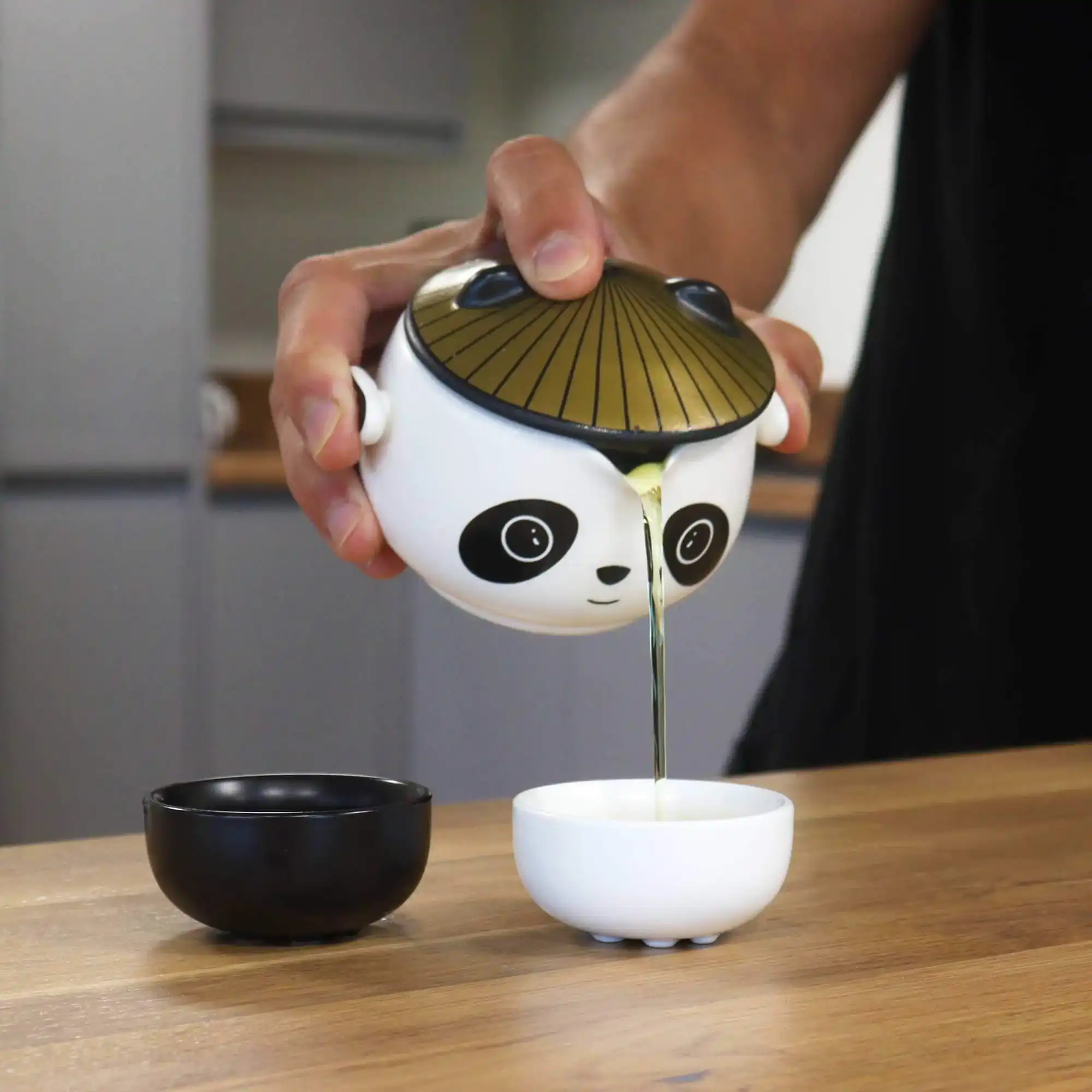A person pours liquid from a panda-shaped container into a small white bowl on a wooden table, with a black bowl nearby.