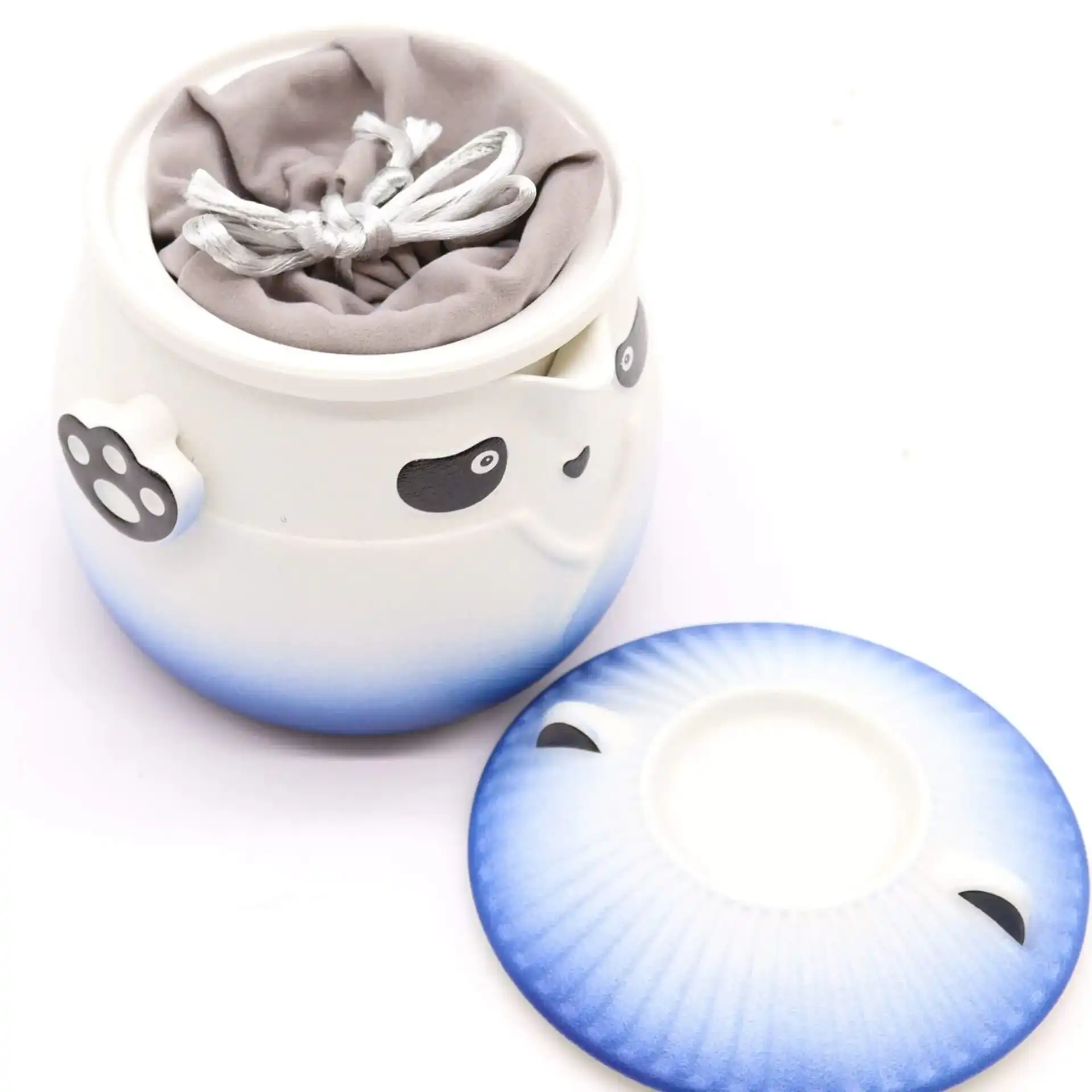 A ceramic container with panda face details and a paw-shaped handle, holding a gray drawstring pouch, with the matching lid placed beside it.