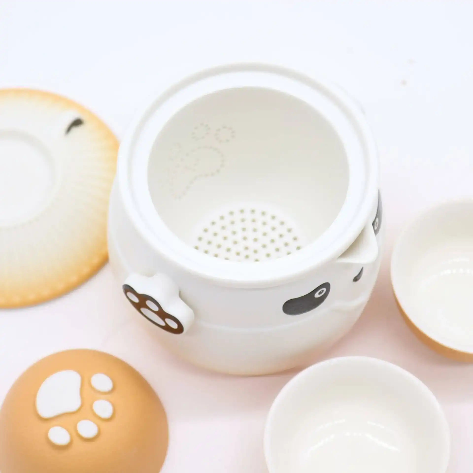 A white ceramic teapot with panda face and paw print designs, accompanied by a matching lid, two small teacups, and a saucer on a white surface.