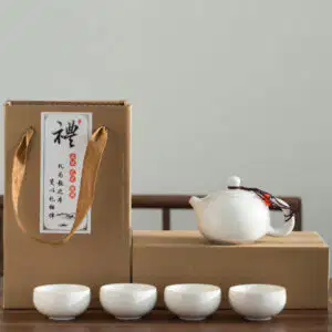 A white ceramic teapot and four matching teacups are displayed in front of a cardboard gift box with Chinese writing and a fabric handle.
