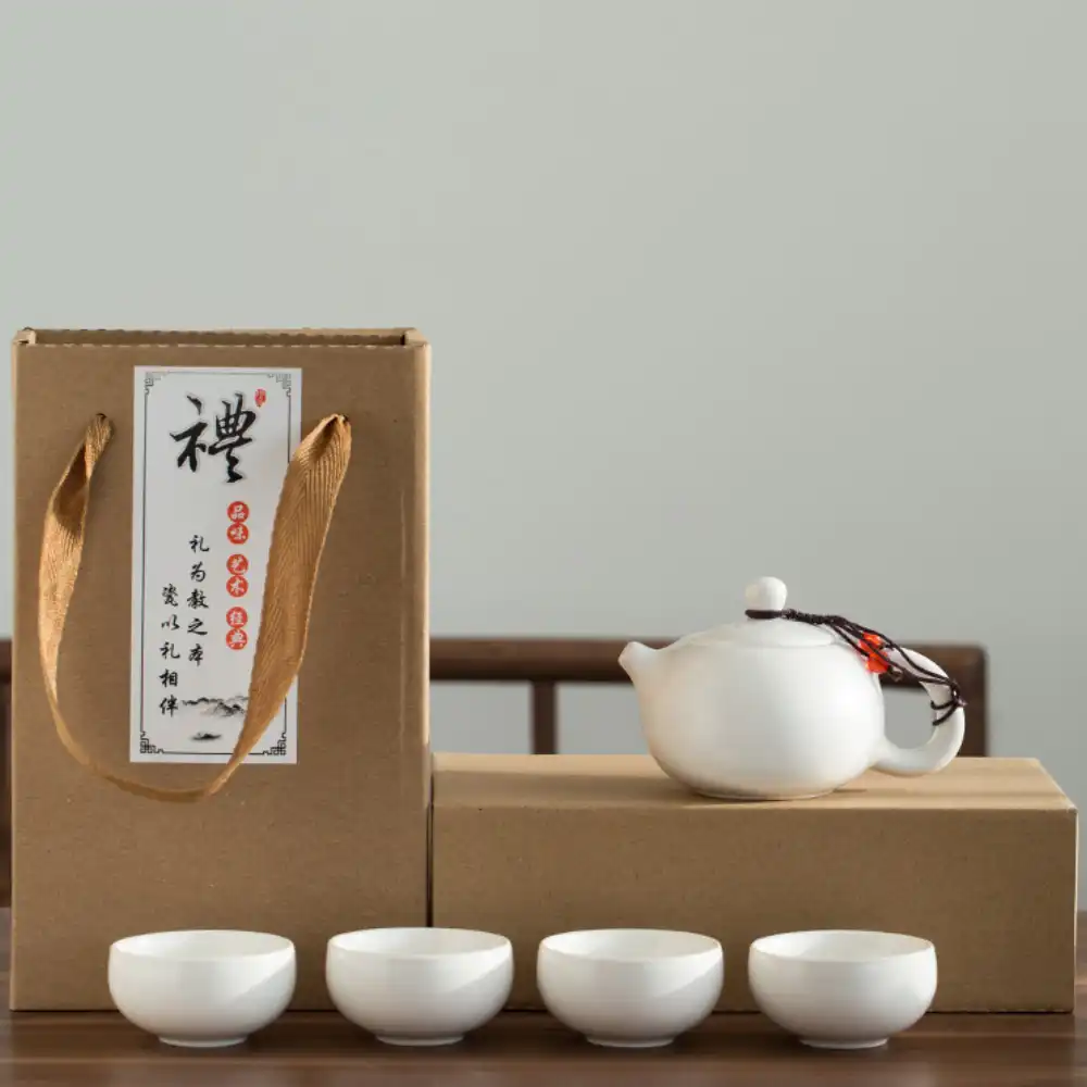 A white ceramic teapot and four matching teacups are displayed in front of a cardboard gift box with Chinese writing and a fabric handle.