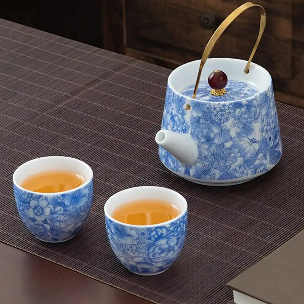 Blue and white floral porcelain teapot with a brass handle next to two matching teacups filled with tea, on a dark striped table mat.