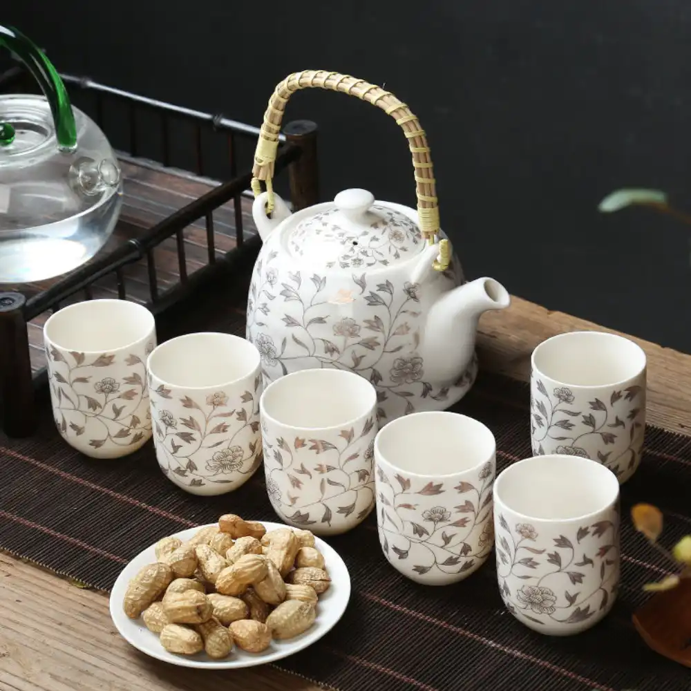 A ceramic teapot and six matching cups with floral patterns are arranged on a mat, alongside a plate of peanuts on a wooden table.