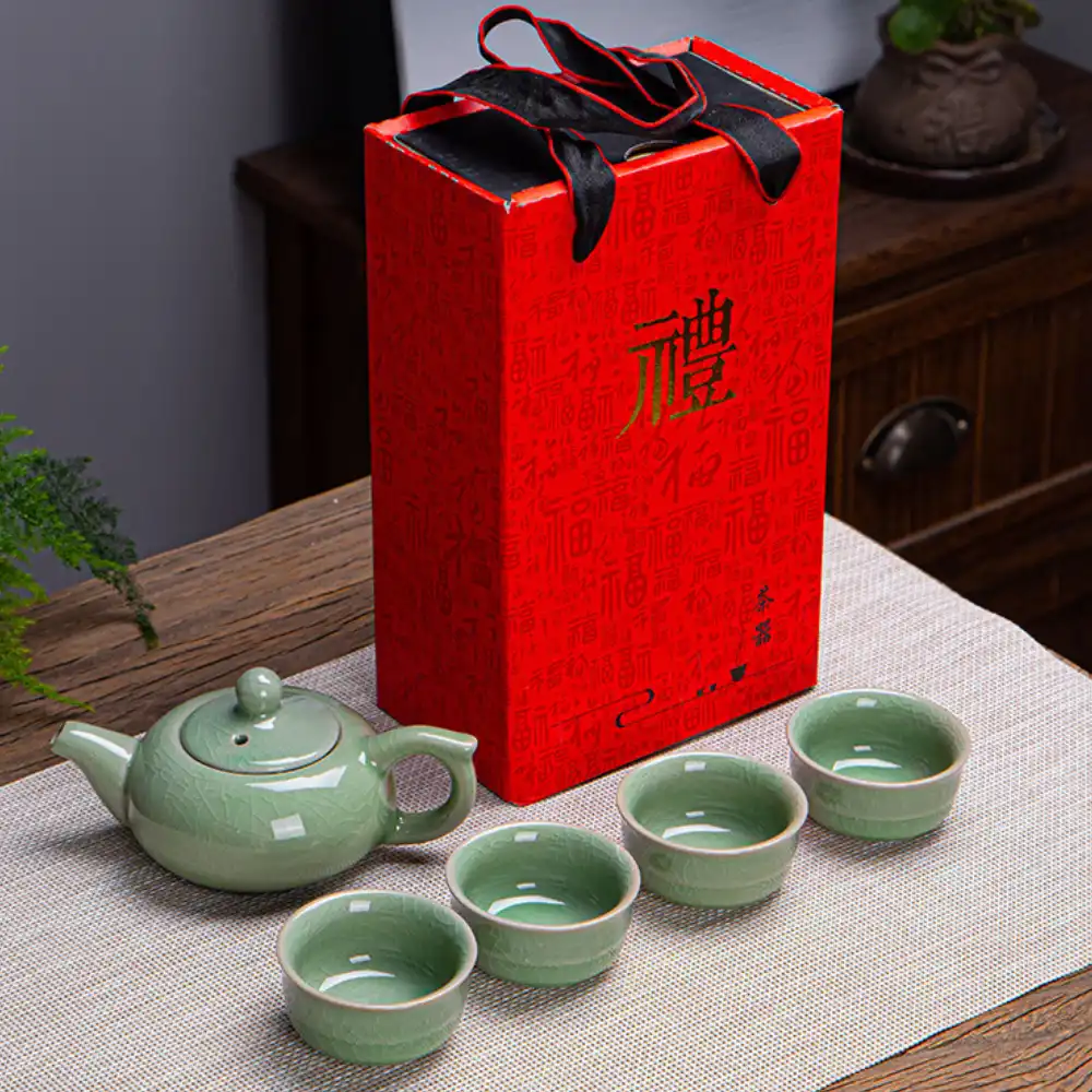 A green ceramic teapot with four matching teacups sits beside a tall red gift box with Chinese characters, placed on a woven mat on a wooden table.