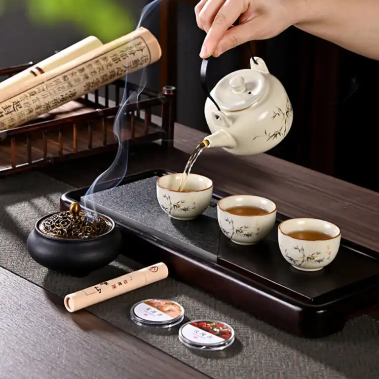 A hand pours tea from a decorated teapot into three matching cups on a tray, with incense burning and scrolls and tea tins nearby on a wooden table.