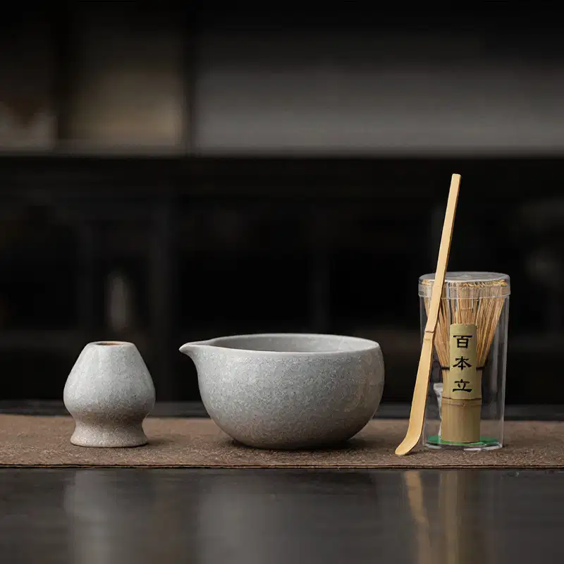 The Kiln-Glazed Matcha Set – 4 Piece | Stone includes a small cup, pouring bowl, bamboo whisk in a clear container, and bamboo scoop on a mat for an authentic tea experience.