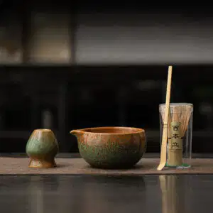 The Kiln-Glazed Matcha Set – 4 Piece | Stone includes a ceramic cup, matching spouted bowl, bamboo whisk, and scoop in a clear container, arranged on a mat against a dark backdrop.