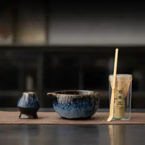The Kiln-Glazed Matcha Set – 4 Piece | Stone, including a ceramic cup, spouted bowl, and bamboo whisk in a clear container, is displayed on a brown mat with a softly blurred stone-toned background.