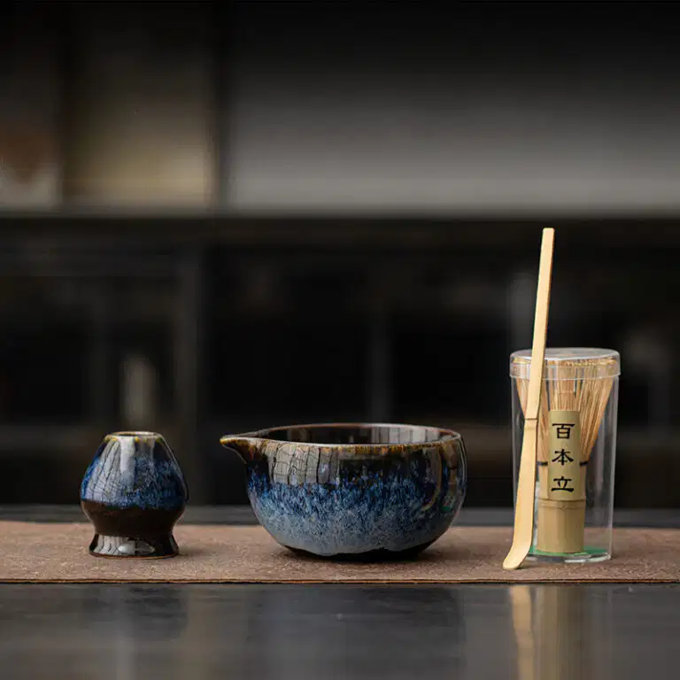 The Kiln-Glazed Matcha Set – 4 Piece | Stone, including a ceramic cup, spouted bowl, and bamboo whisk in a clear container, is displayed on a brown mat with a softly blurred stone-toned background.