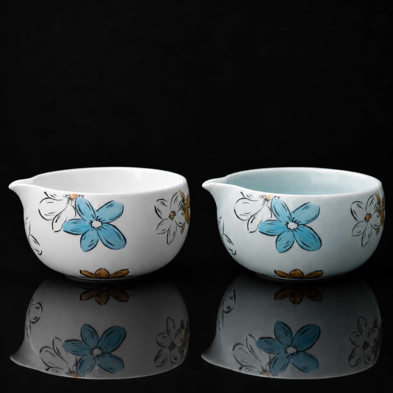 Two small ceramic bowls with spouts, each decorated with blue and yellow flowers reminiscent of a Blossom Matcha set, are placed side by side on a reflective black surface.