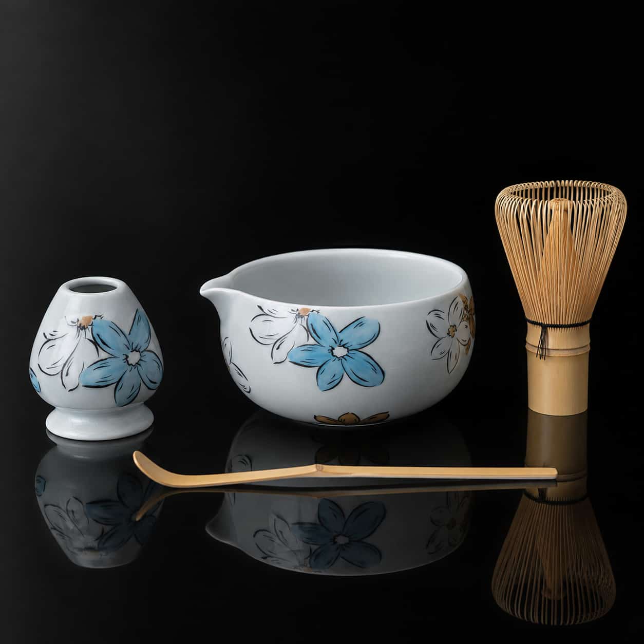 White ceramic Matcha Bowl, whisk holder, bamboo whisk, and bamboo scoop are arranged on a reflective black surface. The ceramics feature blue and yellow floral designs, making this Whisk Gift Set a beautiful addition to any Blossom Matcha ritual.