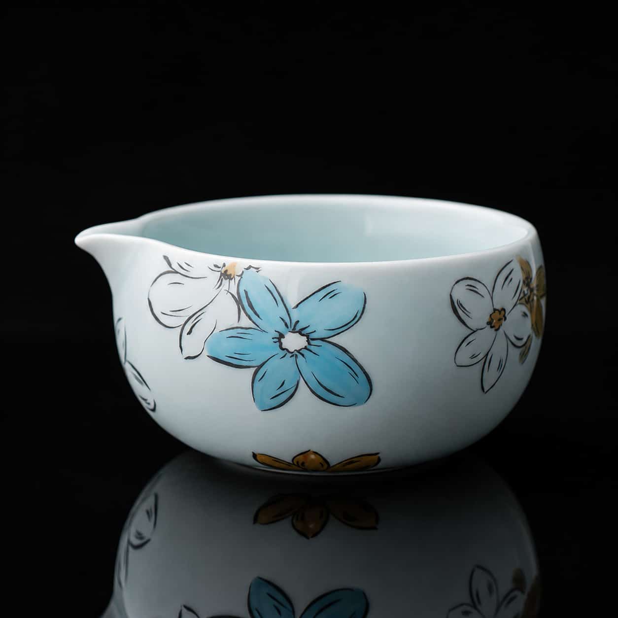 White ceramic Matcha Bowl with spout, decorated with blue and yellow floral patterns, set against a black background with a reflective surface—a perfect addition to any Blossom Matcha Whisk Gift Set.