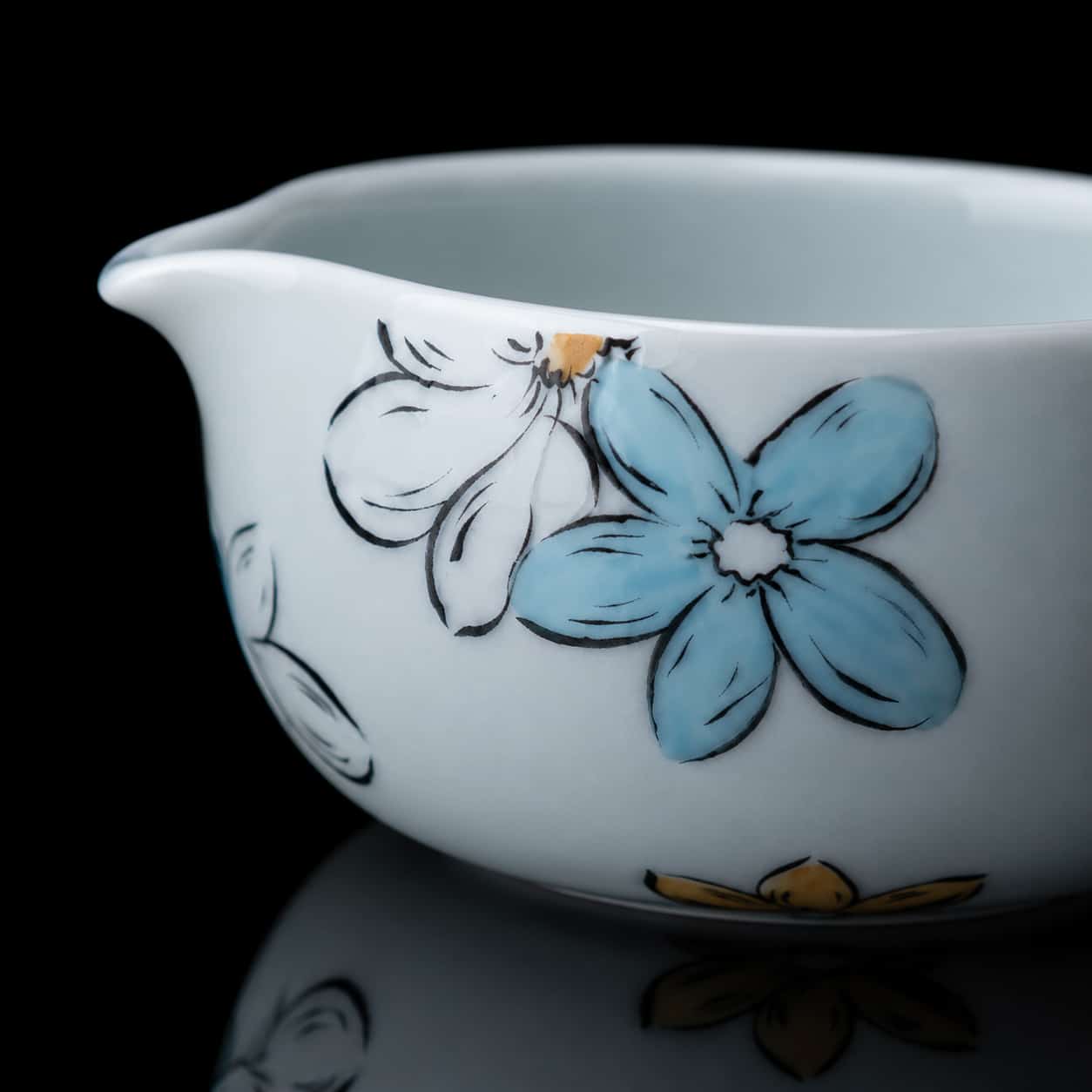 White ceramic Matcha Bowl with a pouring spout, decorated with hand-painted blue and yellow flowers in a delicate Blossom Matcha design, shown on a glossy black surface.