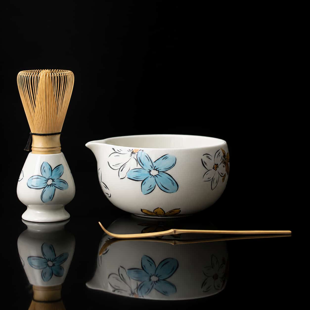 A Blossom Matcha bowl adorned with a delicate floral pattern, a bamboo whisk with a matching holder, and a bamboo tea scoop are artfully arranged on a reflective black surface.