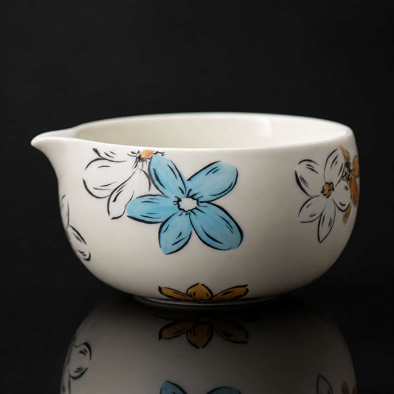 A small, white ceramic matcha bowl with a spout, decorated with blue and yellow floral patterns reminiscent of Blossom Matcha designs, set against a black background.