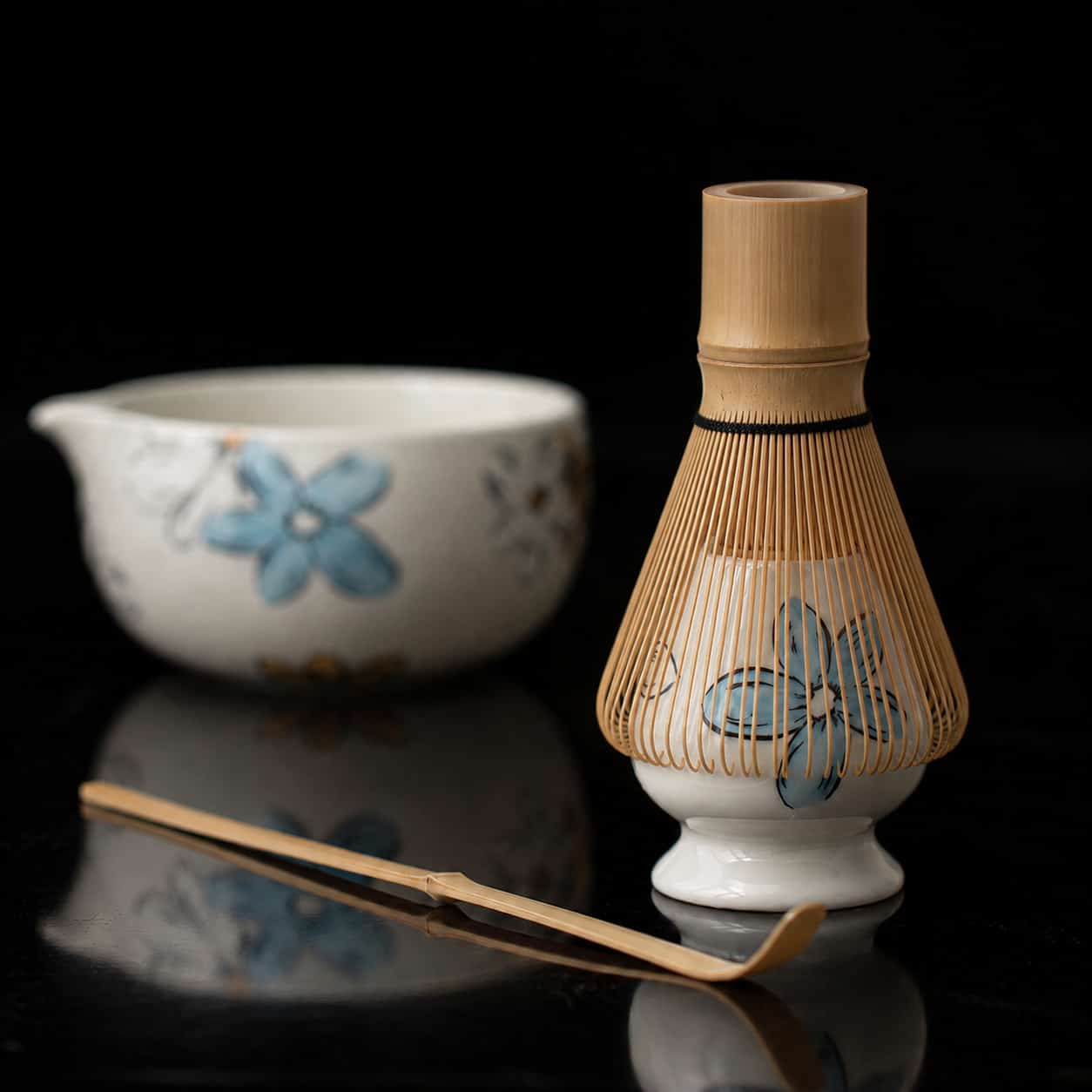 A bamboo matcha whisk, scoop, and a Blossom Matcha bowl with a blue floral design are arranged on a black surface, creating an elegant Whisk Gift Set perfect for any tea lover.