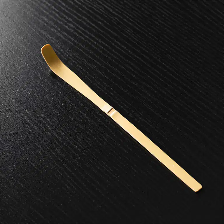 A gold metal tool with a flat handle and curved tip rests on a textured black surface next to the 100-Tine Bamboo Matcha Whisk - Chasen.