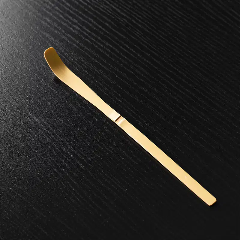 A gold metal tool with a flat handle and curved tip rests on a textured black surface next to the 100-Tine Bamboo Matcha Whisk - Chasen.