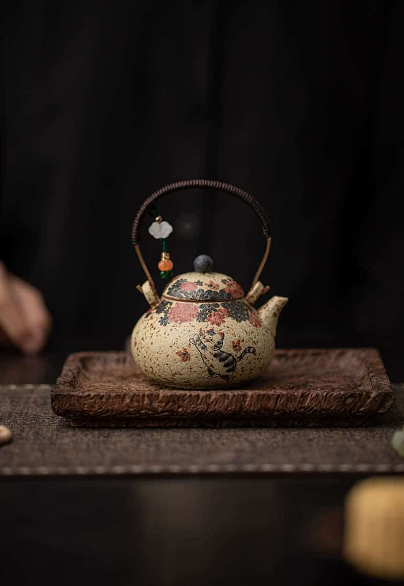 The Whiskers & Blooms Hand-Painted Red Clay Ceramic Teapot-for-One (150ml), adorned with delicate floral designs and a beaded handle, sits on a carved wooden tray against a dark background—making it the perfect teapot for one.