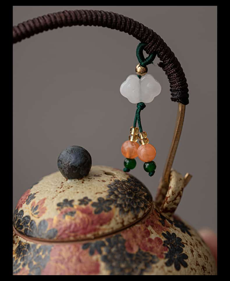 A close-up of the Whiskers & Blooms Hand-Painted Red Clay Ceramic Teapot-for-One’s lid, featuring a floral pattern, a metal handle wrapped in cord, and a beaded charm with white and orange beads.