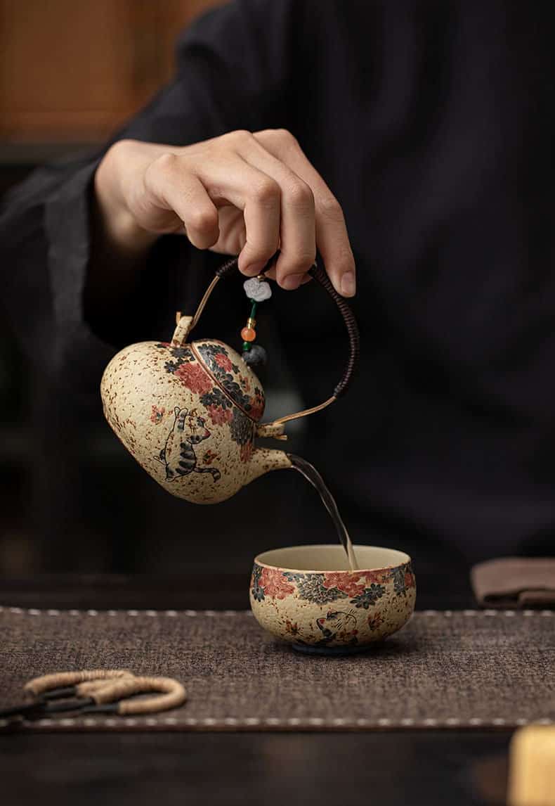 A person pours tea from the Whiskers & Blooms Hand-Painted Red Clay Ceramic Teapot-for-One (150ml) into a matching teacup on a woven placemat.