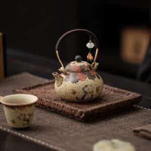 The Whiskers & Blooms Hand-Painted Red Clay Ceramic Teapot-for-One (150ml) with matching cup rests on a textured tray atop a dark table, showcasing elegant floral patterns and a beaded ornament accenting the handle.