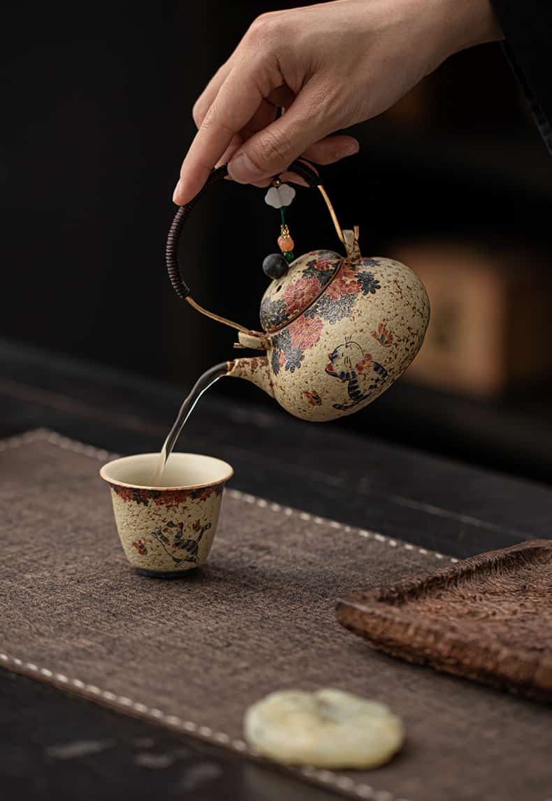 A hand pours tea from the Whiskers & Blooms Hand-Painted Red Clay Ceramic Teapot-For-One (150ml) into a matching teacup placed on a brown mat, with a woven tray and a round stone nearby.